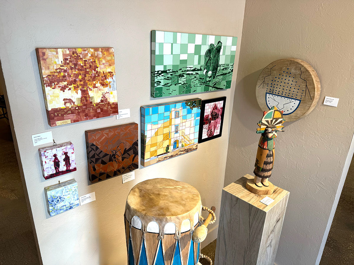 Corner of gallery displaying paintings on wall, sculpture on stand and drum on the floor