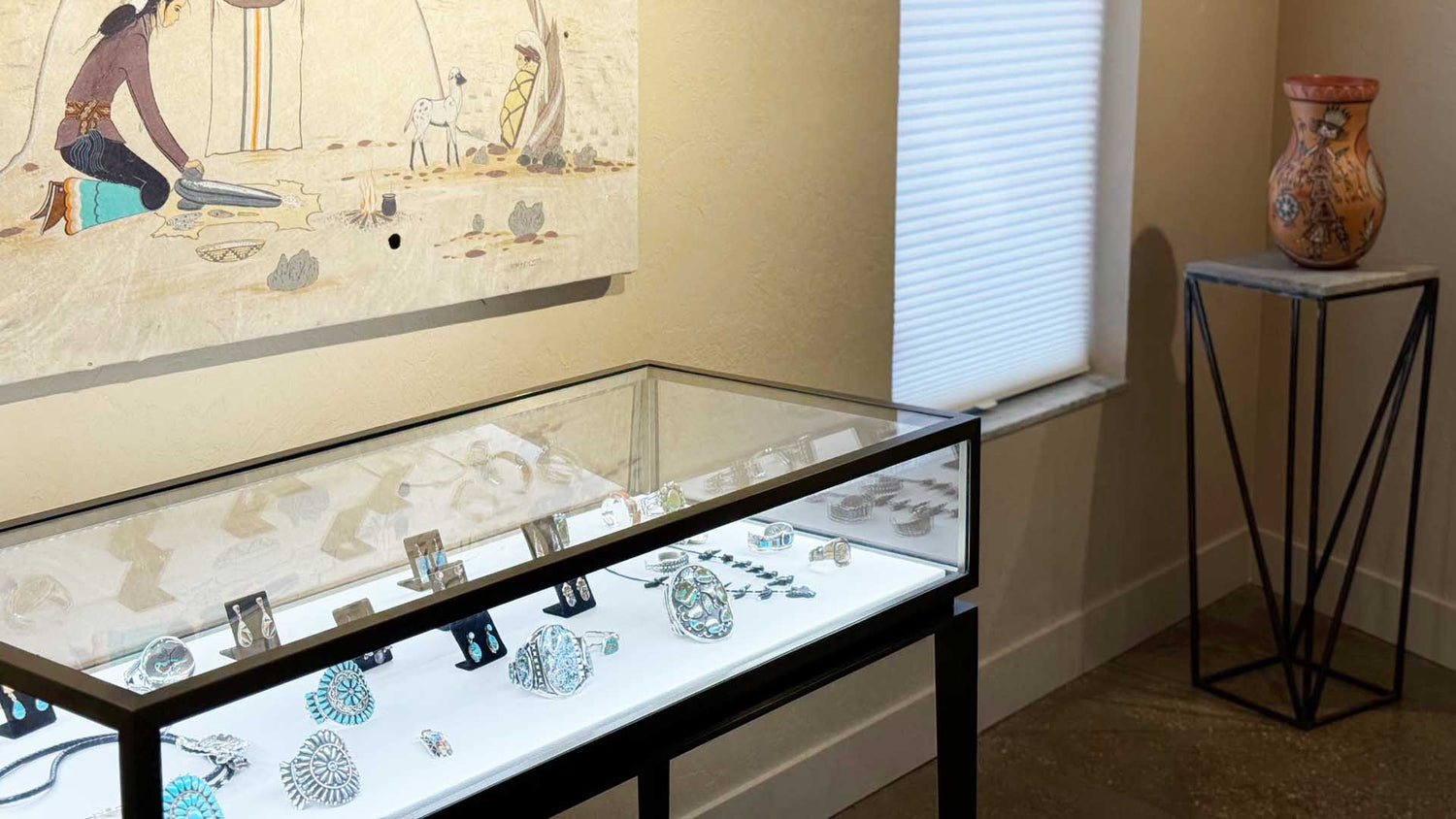 Display case with jewelry in a room with a painting on the wall and a vase on a stand.